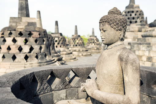 (Selective focus) Stunning view of a Buddha Statue in the foreground and some bell shaped stupas in the background. Borobudur is a Mahayana Buddhist temple in Indonesia.