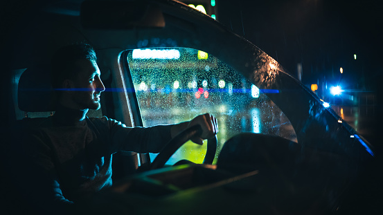 The young man sits in the car on the night rainy street
