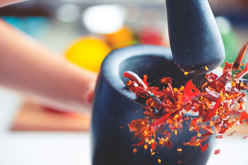 Action image of dried chilli peppers being crushed in a mortar and pestle. The chillis are flying out of the bowl as they are being smashed. Chillis are mostly crushed into flakes