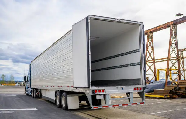 Photo of Big rig blue semi truck with open door semi trailer standing at warehouse parking lot at industrial area waiting for the load for the next delivery