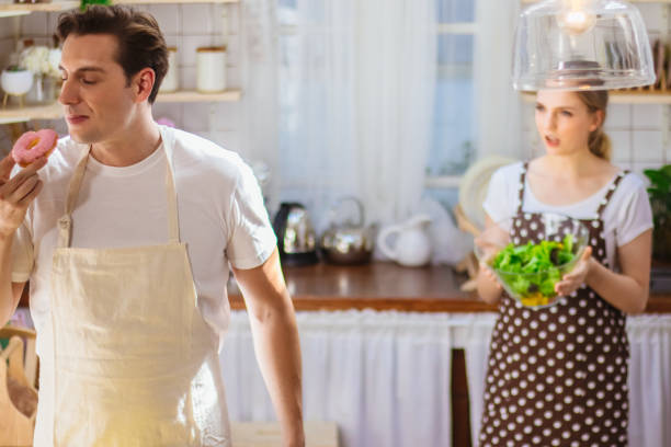 beau couple caucasien ou amant dans la cuisine ensemble. l’homme heureux mangent la malbouffe malsaine comme donut tandis que la salade de cuisson de femme pour le repas. petite amie malheureuse bouleversée ou déçue sur de mauvaises habitudes de petit - overweight women salad frustration photos et images de collection