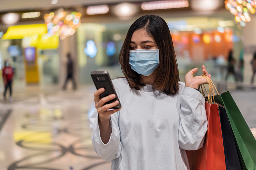 young woman using smartphone at shopping mall and her wearing medical mask for prevention from coronavirus (Covid-19) pandemic. new normal concepts