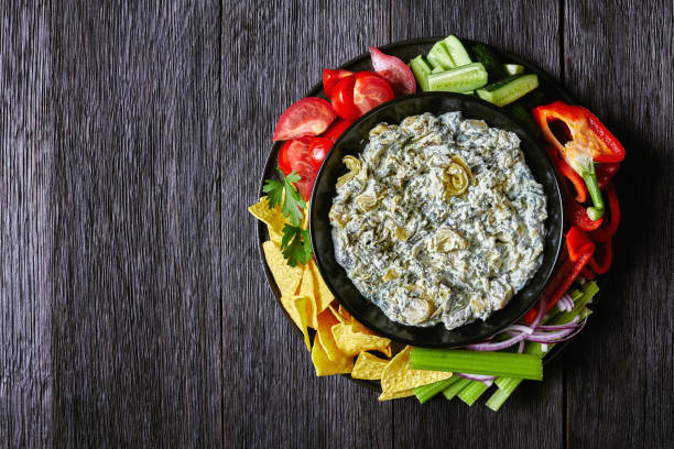 Spinach Artichoke leafy cheese Dip in a bowl with Tortilla Chips, cucumber, celery sticks, tomato slices and red bell pepper, horizontal view from above, flat lay, free space