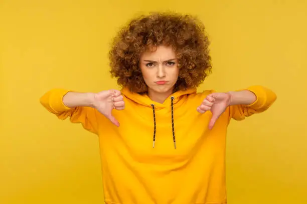Dislike! Portrait of displeased frustrated curly-haired woman in urban style hoodie showing thumbs down, criticizing dissatisfied with awful service. indoor studio shot isolated on yellow background