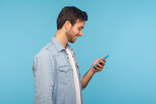 Side view of happy smiling man in worker denim shirt texting in social media, reading positive message on cell phone, using mobile network services. indoor studio shot isolated on blue background