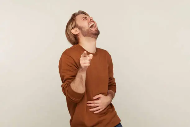 Photo of Hey you! Portrait of amused man with beard in sweatshirt holding hand on stomach and laughing out loud