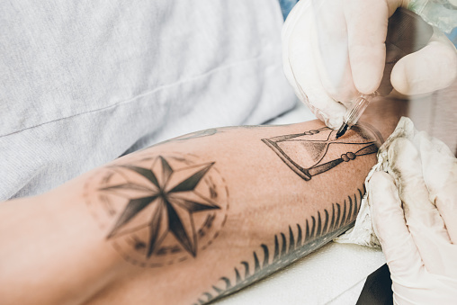 Tattoo artist with needle in his hand, drawing a hourglass tattoo on a man's arm.