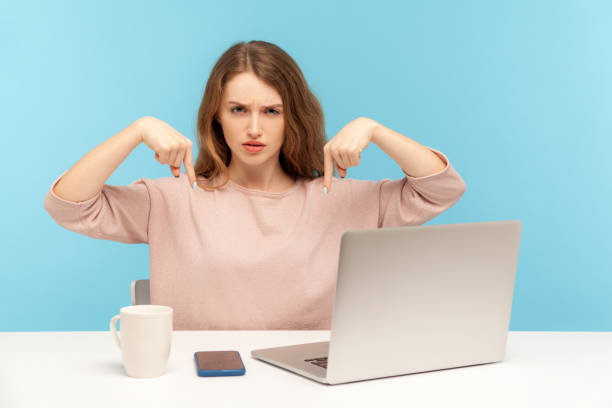 istock Here and right now! Young strict lady boss sitting at workplace with laptop and pointing fingers down, controlling 1255149684