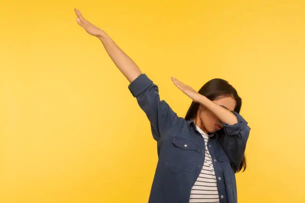 Photo of Yes, success! Portrait of lucky triumphal girl in denim shirt raising hands, moving in dab dance, celebrating victory
