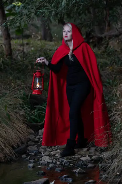 Attractive woman dressed a little red riding-hood cross a stream in a dark forest with a lantern