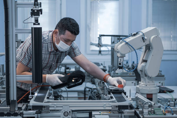 istock industry 4.0 : Man with surgical mask and gloves against COVID-19 ( Coronavirus ) programming robot arm with teach pendant. 1255152271