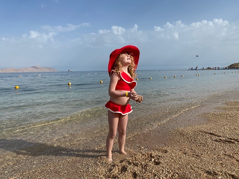 The child is resting in Egypt. A little girl in a red swimsuit and a sun-protection hat stands on the shore of the Red Sea.