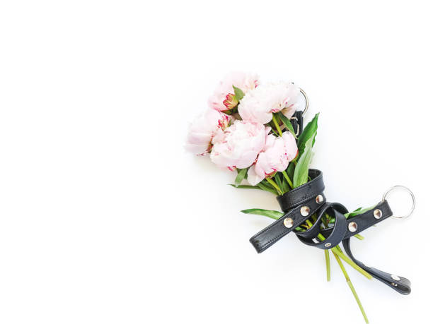 A bouquet of pink peonies is pulled together with a black leather belt. White background. Layout for fans of BDSM, sex shops. Flat lay. Copy space. stock photo