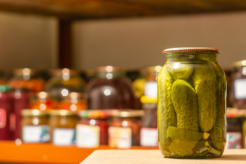 Homemade glass jar with pickled cucumbers in the cellar on the background of shelves with canned vegetables and fruits. Closeup, selective focus