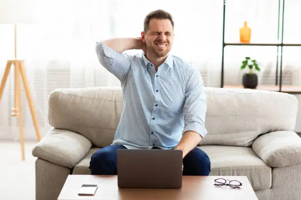 Young frustrated man having strong pain in neck and massaging it, sitting on the couch at desk