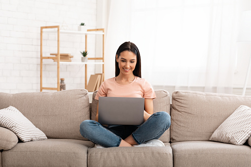 Frontal portrait of happy girl chatting online on laptop with friend at home, sitting on beige couch in light living room