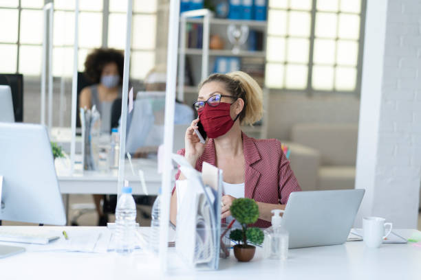 Businesswoman speaking on phone wearing face mask during Coronavirus pandemic. Business colleagues working at office by maintaining social distance due to Coronavirus pandemic. Office with acrylic glass partition on desk office cubicle mask stock pictures, royalty-free photos & images