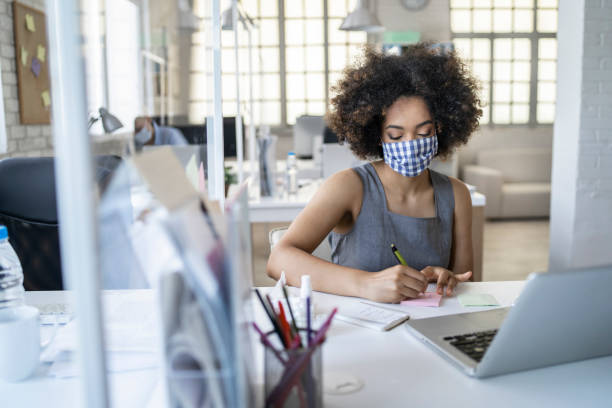 The new normal: Working at office wearing protective face masks maintaining social distance. Businesswoman wearing face mask working at office by maintaining social distance due to Coronavirus pandemic. Office with acrylic glass partition on desk office cubicle mask stock pictures, royalty-free photos & images