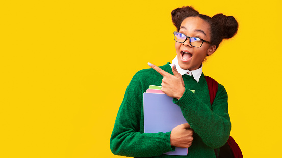 Great Offer. Excited afro teenage girl in glasses pointing at copyspace, holding folders, wearing backpack, panorama