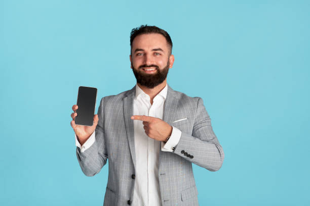 istock Happy office worker pointing at mobile phone with blank screen on blue background, space for design 1255173397