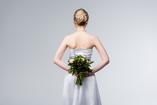 back view of bride in elegant wedding dress holding bouquet of flowers behind back isolated on grey