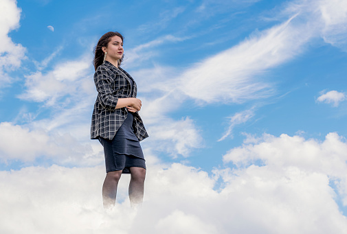 A young girl slowly walks through the clouds in a dark dress and a checkered jacket on a background of blue sky