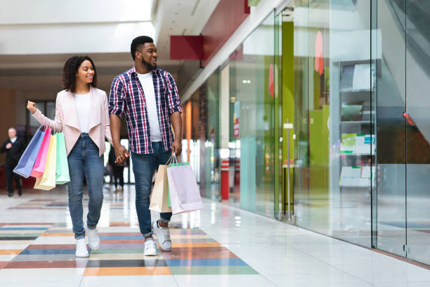 Seasonal Sales. Happy Black Couple Walking In Mall Center After Buying Clothes Seasonal Sales. Happy Black Couple Walking In Mall Center After Buying Clothes, Looking At Store Window, Copy Space mall walker stock pictures, royalty-free photos & images