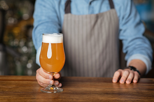 Fresh craft beer. Man in apron puts on wooden table glass of light ale with foam, close up, cropped
