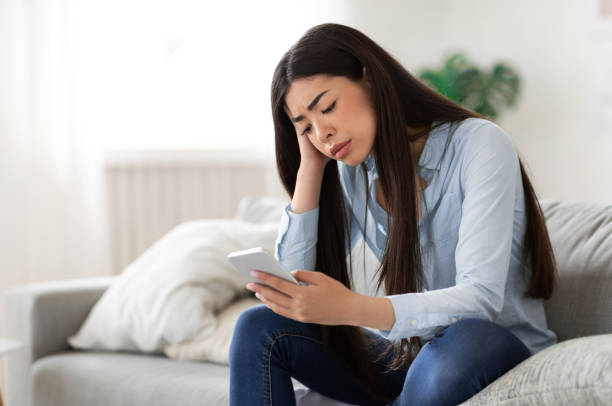 Waiting for his call. Upset asian girl sitting with cellphone at home Waiting for his call. Upset asian girl looking sadly at mobile phone screen, sitting on sofa at home, copy space co dependent relationship stock pictures, royalty-free photos & images