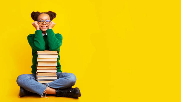 Afro schoolgirl with stack of books at studio Bookworm. Dreamy afro teenage girl sitting on floor with pile of books over yellow background, copyspace, panorama book bookstore sale shopping stock pictures, royalty-free photos & images