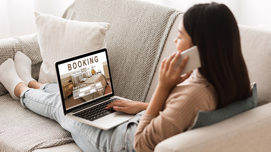 Asian girl booking hotel online, talking on phone with reservation team, sitting on couch at home at using laptop