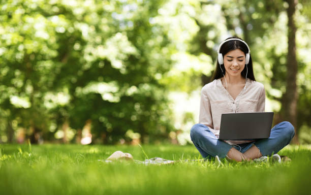 menina asiática alegre sentada no gramado com laptop, estudando e ouvindo música - laptop computer grass nature - fotografias e filmes do acervo