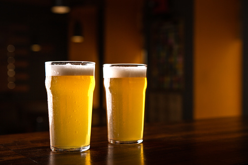 Order beer at pub. Two glasses of light ale on table in interior of dark pub, close up, free space
