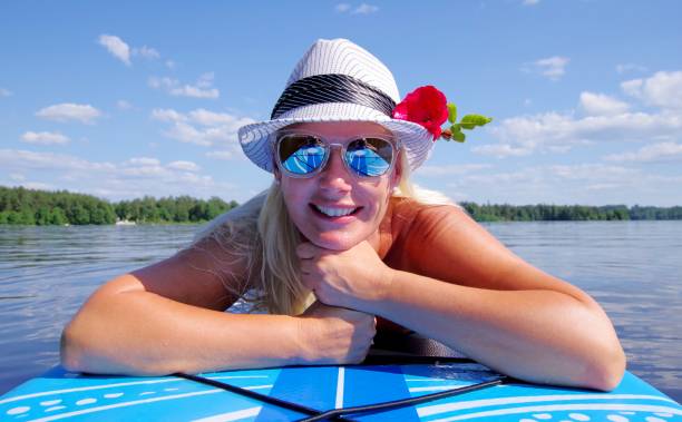 mulher loira feliz com chapéu branco com rosa, deitada no paddleboard, céu e água ao fundo. - sweden summer swimming lake - fotografias e filmes do acervo