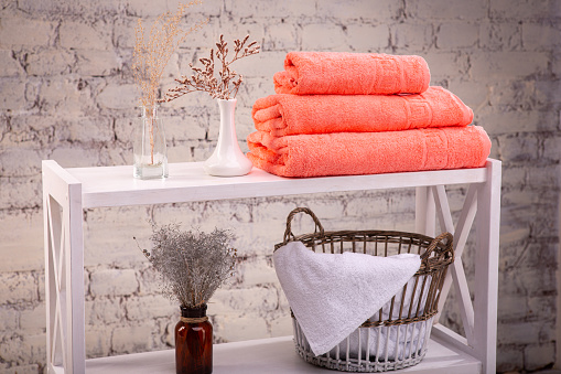 Rack with a stack of three peach color towels and baskets with clean white towels and toilet decor near a brick wall. Shelf with terry towels and decorative vases. Shelving unit with clean towels.