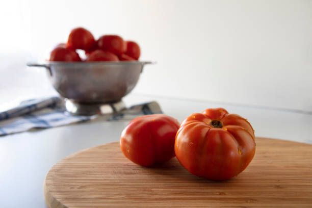 Close up of Fresh Ripe Tomatoes on a Cutting Board and Behind Tomatoes in a Colander on White Background