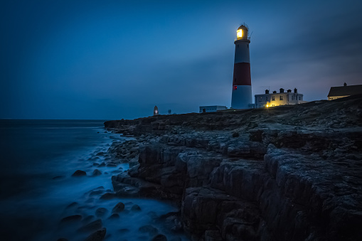 View of Portland Bill Lighthouse at night, with waves going over rocks in the foreground. Dorset, UK