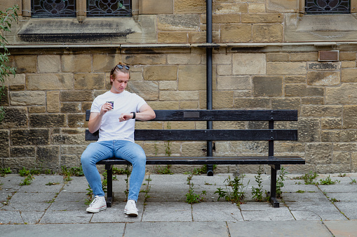 A young man wearing casual clothing is sitting down on a bench outdoors, he is looking at his smartwatch and holding a coffee cup while waiting for a friend.