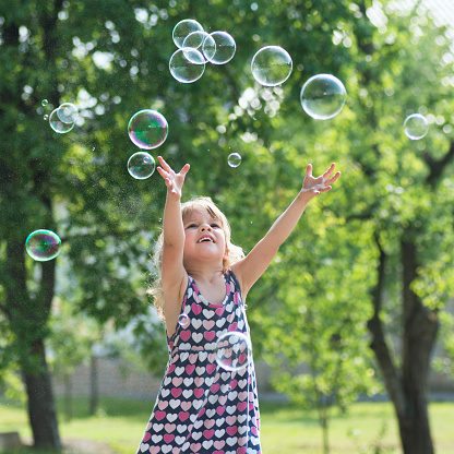 The cute little girl in dress is playing with soap bubbles outdoors.