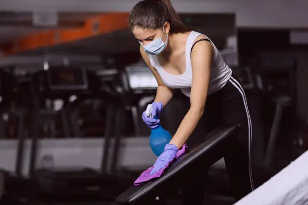 Female gym staff spraying and wiping equipment with sanitizer in order to prevent coronavirus infection. COVID-19 concept. Woman wearing surgical gloves and mask in fitness club.
