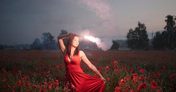 Portrait of pretty brunette girl in a red dress dancing with red burning signal flare in poppy field.