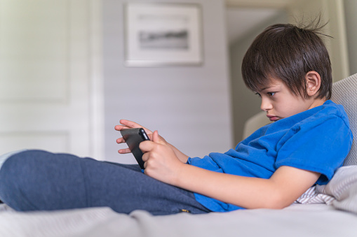 An ethnic nine-year old boy plays a game on a digital tablet. He is sitting on a couch in a modern living room.
