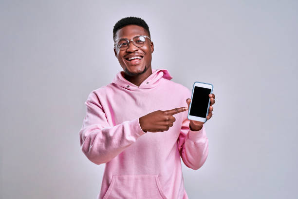 alegre hombre afroamericano vestido con una sudadera rosa con capucha y gafas sostiene un teléfono y muestra en su pantalla. el concepto de gadgets y anuncios. - afro man fotografías e imágenes de stock