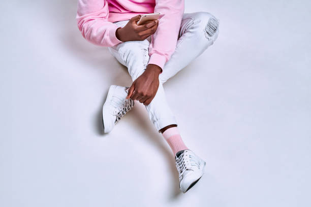 scatto ritagliato di un ragazzo vestito con una felpa rosa con cappuccio, pantaloni bianchi, calze rosa e scarpe da ginnastica bianche tiene un telefono. il concetto di gadget, accessori, abbigliamento e lifestyle. - modella negozio foto e immagini stock