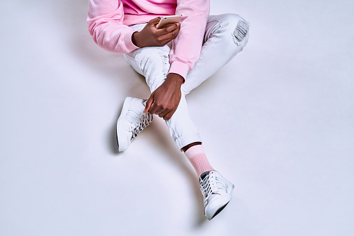 Cropped shot of a guy who dressed in a pink hoodie, white pants, pink socks and white sneakers holds a phone. The concept of gadgets, accessories, clothing and lifestyle.