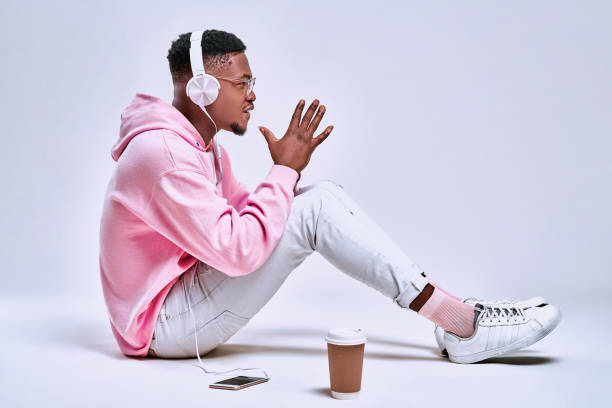 Stylish african american guy dressed in a pink hoodie,white pants, pink socks,glasses, listens to music in white headphones and drinks a coffe. The concept of gadgets, accessories, clothing,lifestyle. Stylish african american guy dressed in a pink hoodie,white pants, pink socks,glasses, listens to music in white headphones and drinks a coffe. The concept of gadgets, accessories, clothing,lifestyle. running shoes on floor stock pictures, royalty-free photos & images