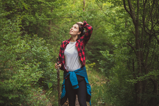 Closeup view of a woman fully equipped for a day in nature. She is walking in the forest with a big stick in her hand and looking around.