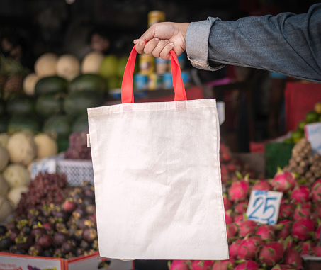 hands holds a white cloth bag in local fruit market as background. concept for slow life, sustainable style, renewable , reduce plastic use.