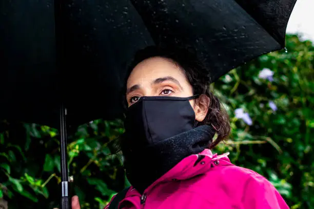 a mature woman in a pink coat, holding umbrella on the street and wearing a black mask due to the covid-19