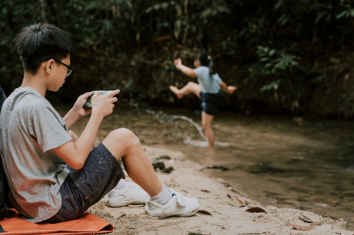 an asian chinese boy sitting alone at the riverside playing mobile games with his smart phone while his sister playing in the river in the forest during weekend